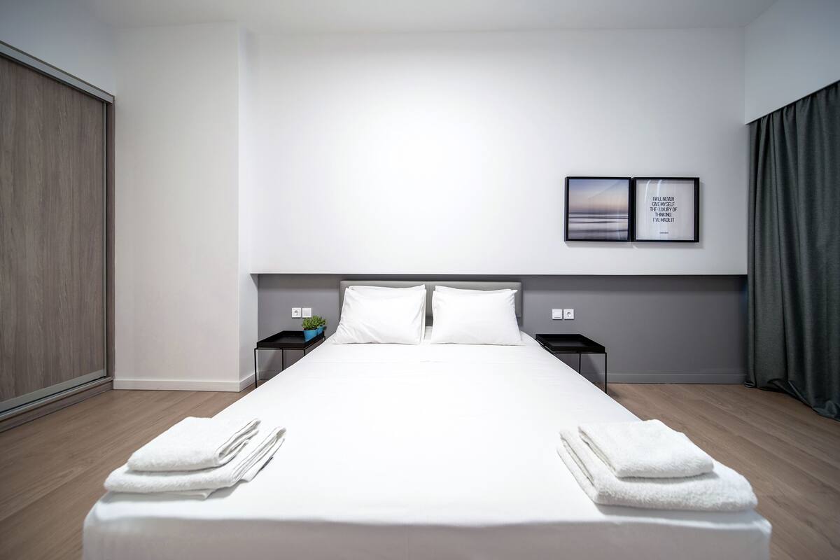 Quarto de hotel moderno com uma cama grande, duas mesas de cabeceira, um criado-mudo, TV fixada na parede e vista para a cidade.