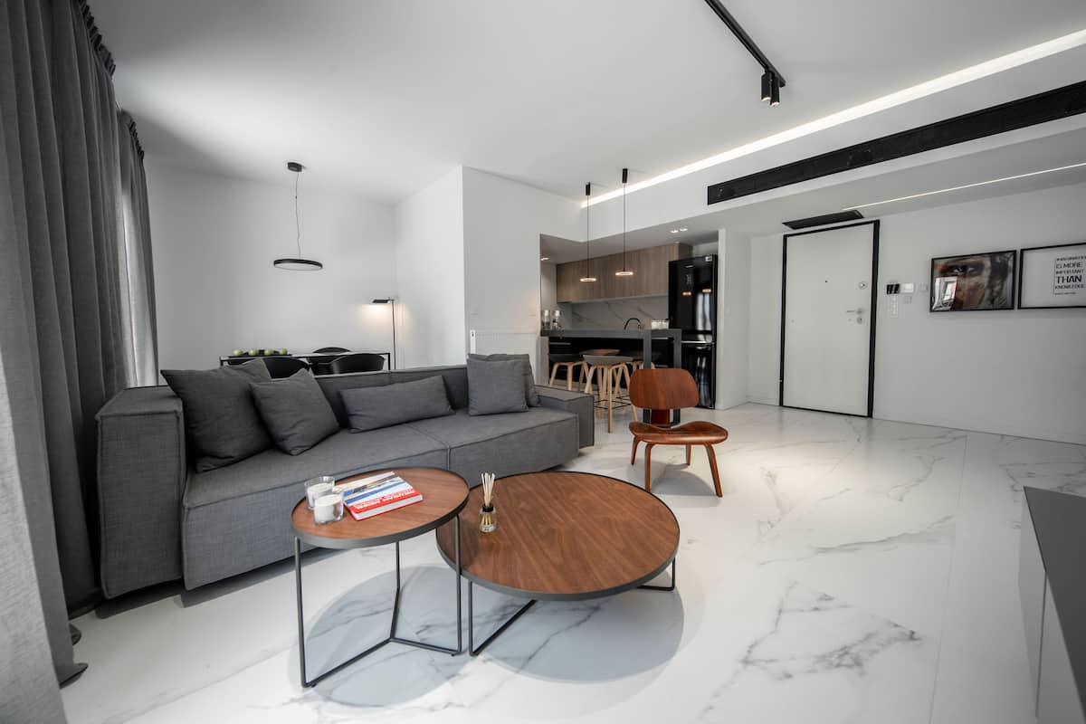Uma sala de estar moderna com um sofá cinza, duas mesas de centro e uma cozinha ao fundo.