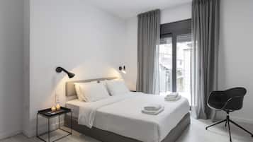 Habitación de hotel moderna con una cama grande, una mesita de noche con lámpara, una silla y vistas a edificios a través de la ventana.