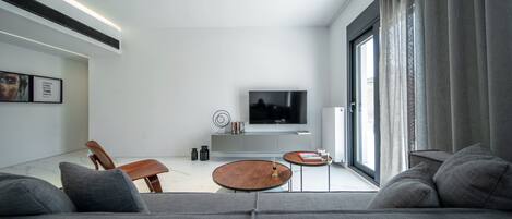 A modern living room with a grey sofa, a wooden coffee table, a TV on a white stand, and a large window with curtains.