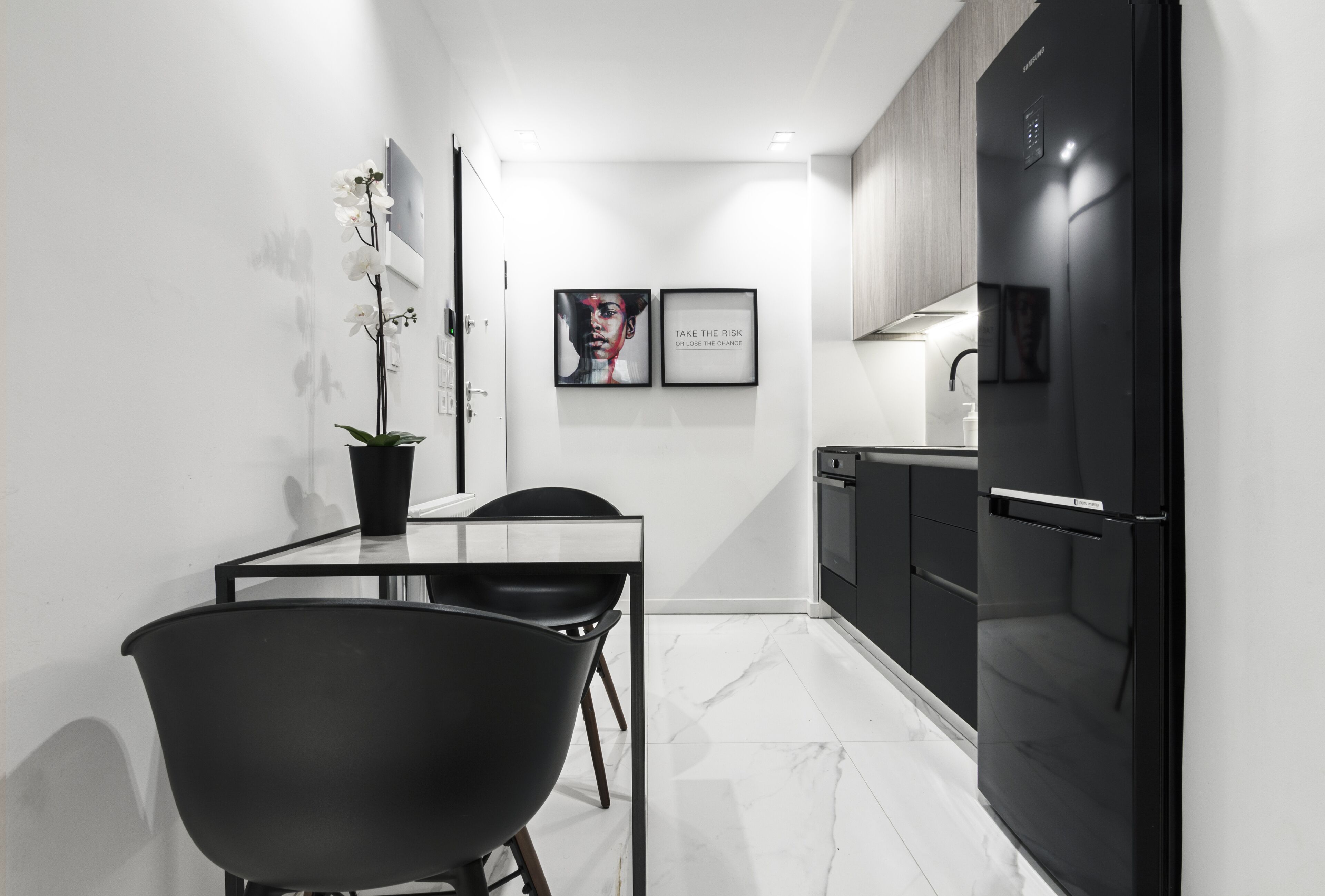 Une cuisine moderne avec des meubles de rangement noirs, un réfrigérateur noir et un coin repas comprenant une table et des chaises. Deux tableaux encadrés ornent le mur.