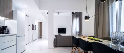 A modern kitchen with white cabinets and a marble floor, leading to a dining area with a table, chairs, and a TV mounted on the wall.