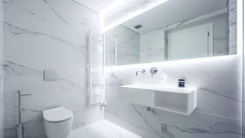 Une salle de bain moderne avec un sol et des murs en marbre blanc, des toilettes blanches, un lavabo encastré avec un miroir au-dessus et un porte-serviettes.