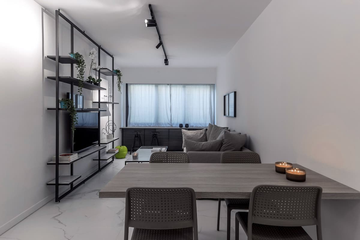 Uma sala de estar moderna com área de jantar, apresentando uma janela grande, um sofá, uma televisão e uma estante com plantas.