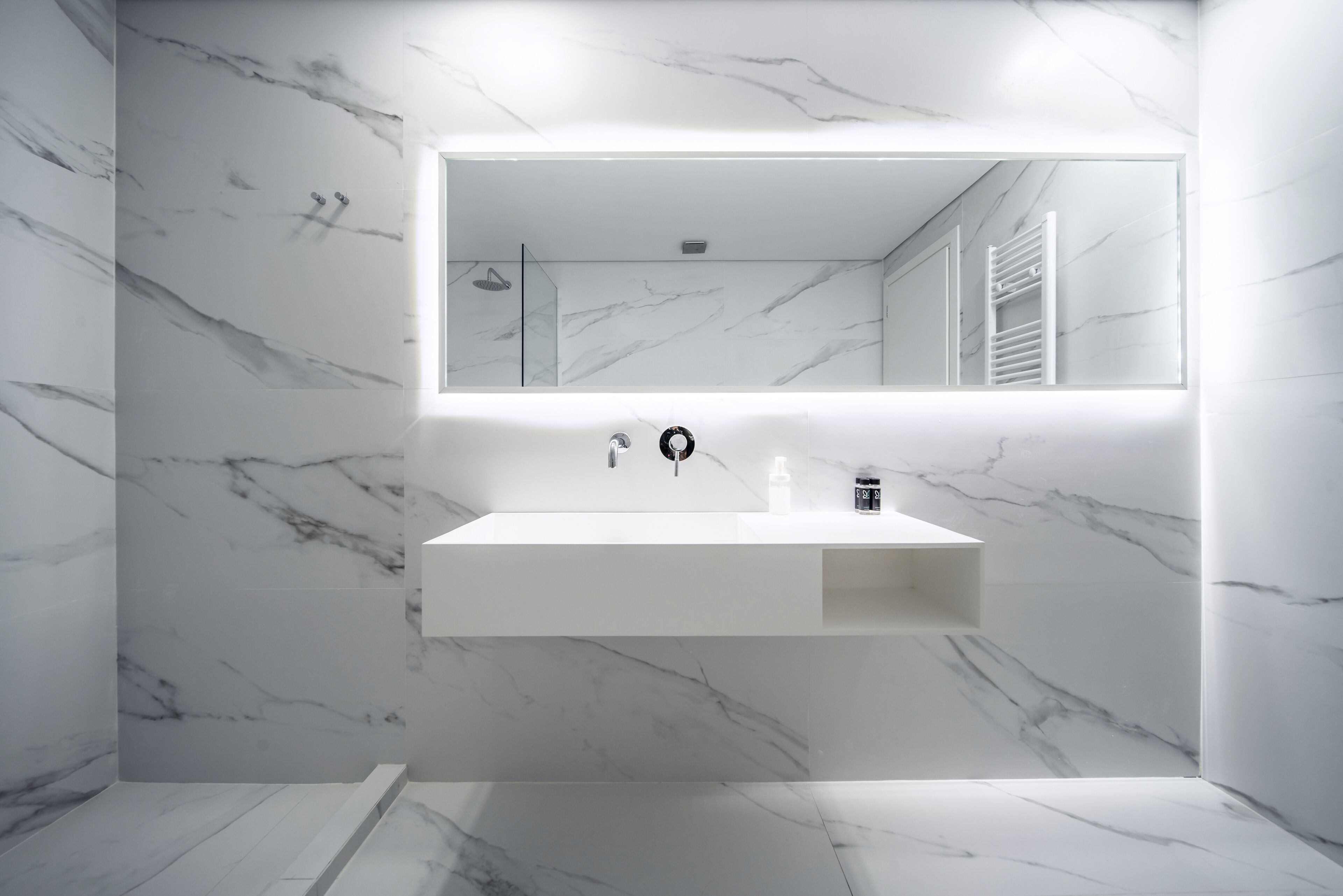 A modern bathroom with a marble wall, a large mirror, a white sink, and a shower area.