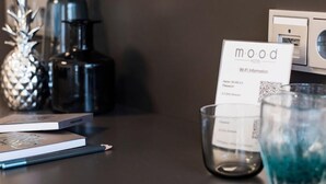 Habitación de hotel con diseño moderno, que incluye una mesa de madera oscura con un libro, una vela y un vaso con líquido azul. Se ve un cartel con información de Wi-Fi.