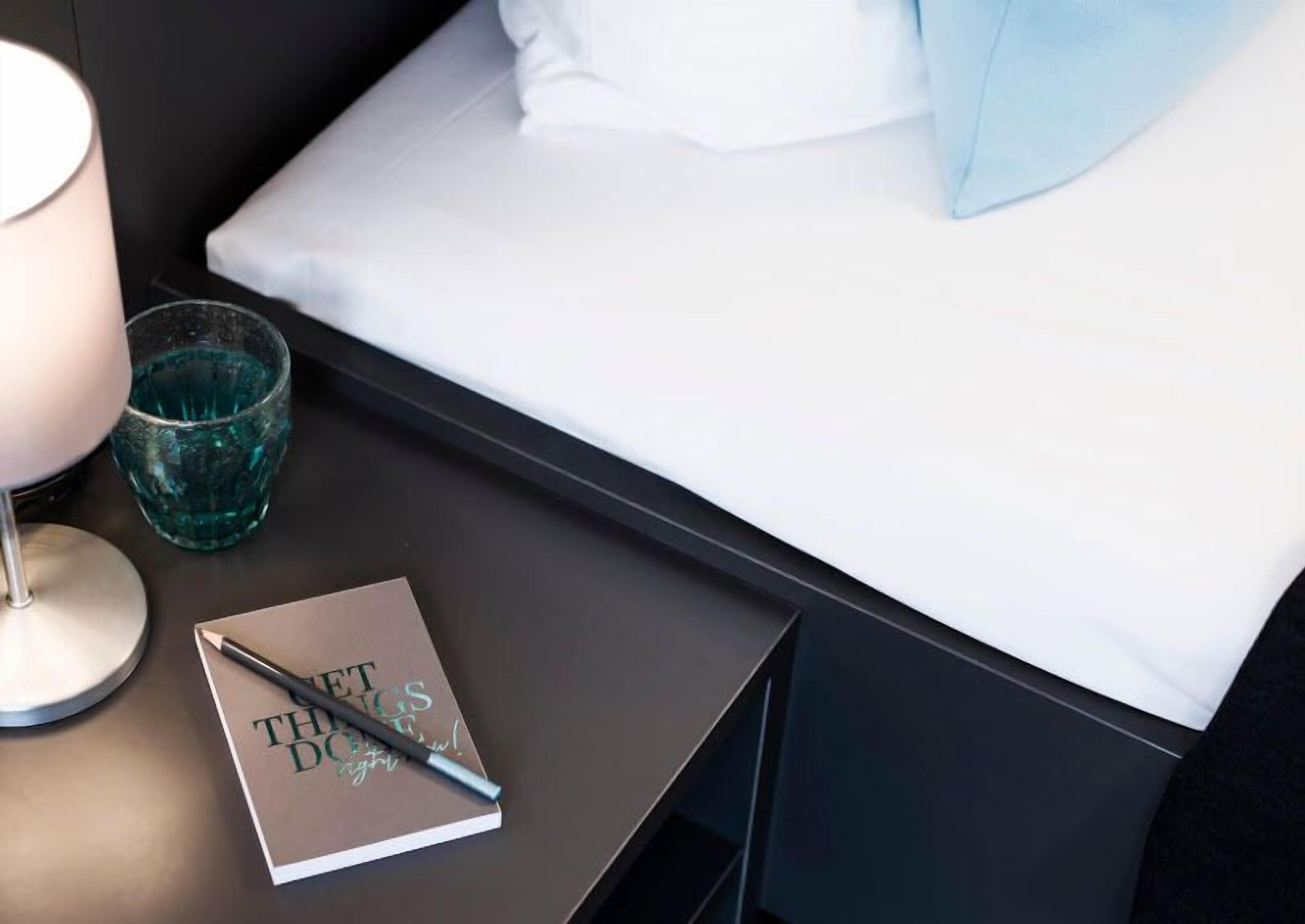 Ein ordentlich gemachtes Bett mit weißer Bettwäsche und einem hellblauen Kissen, ein Nachttisch mit einer Lampe und einem Glas Wasser, sowie ein Notizbuch mit dem Schriftzug 