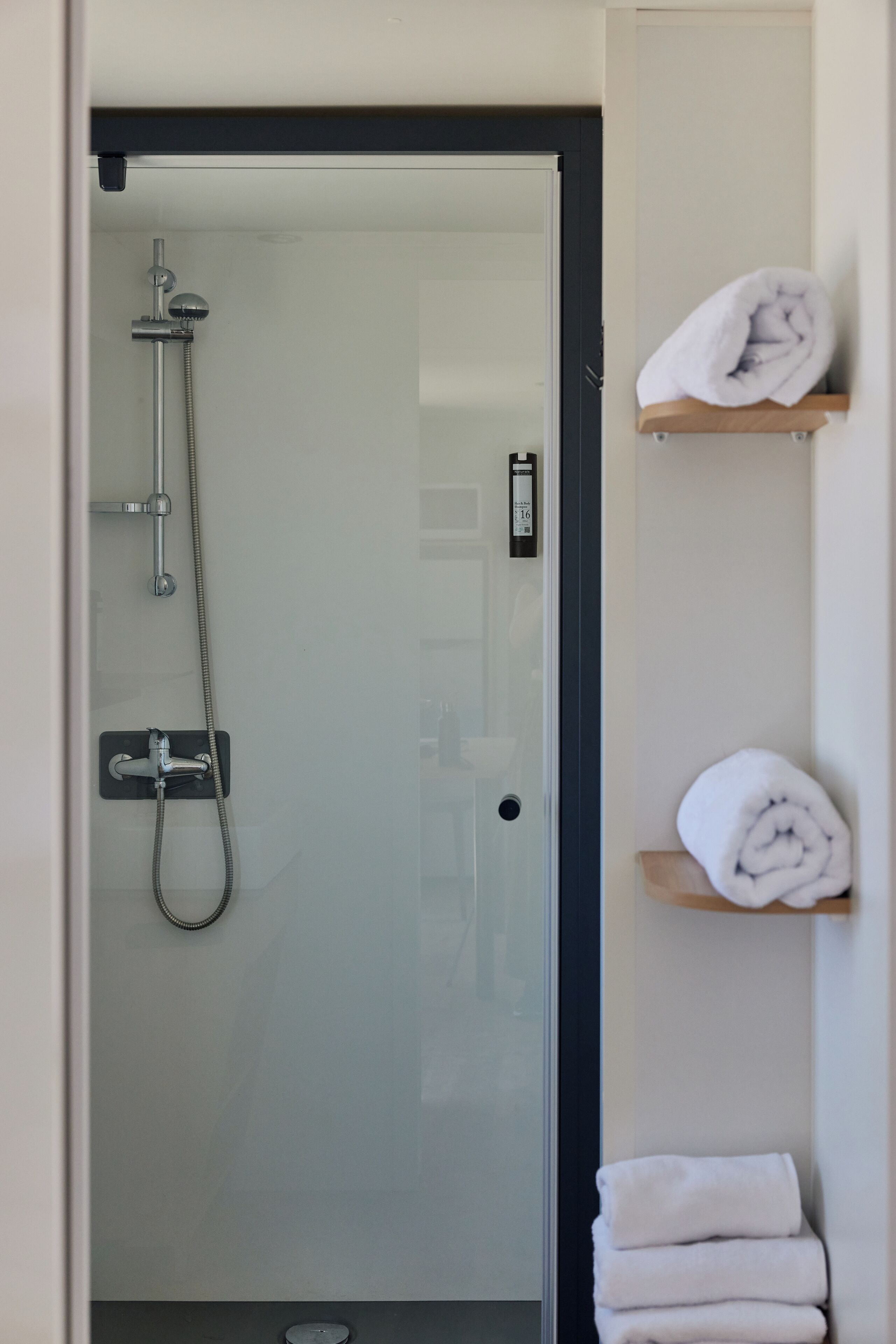 A glass-enclosed shower area with a handheld showerhead and a wall-mounted faucet. There are two white towels stacked on a wooden shelf to the right.
