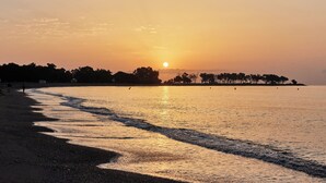 Una playa al atardecer con un mar tranquilo, cielo despejado y siluetas de árboles y gente.