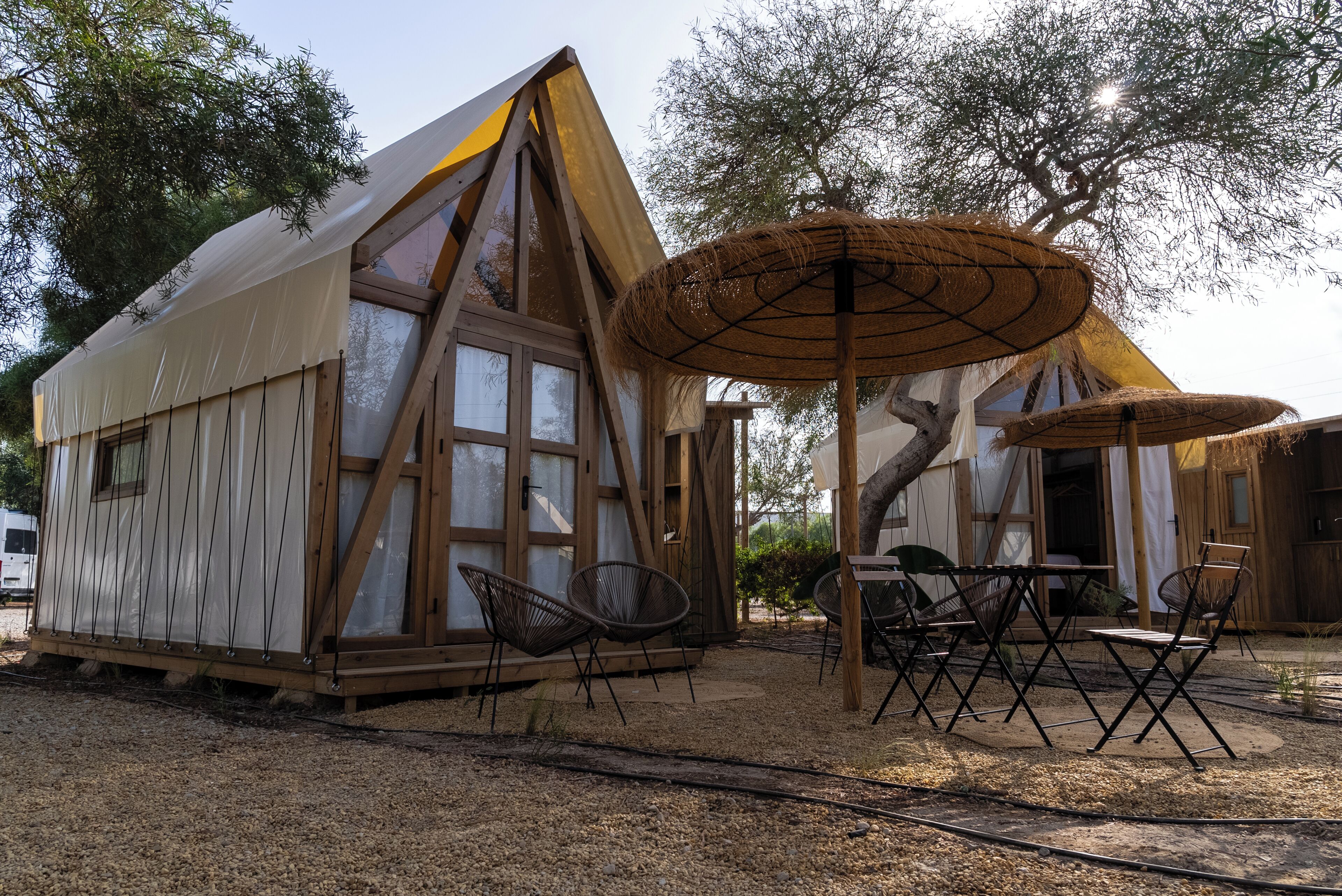 A small, wooden cabin with large windows, a dining area with chairs and a table, and a thatched roof structure.