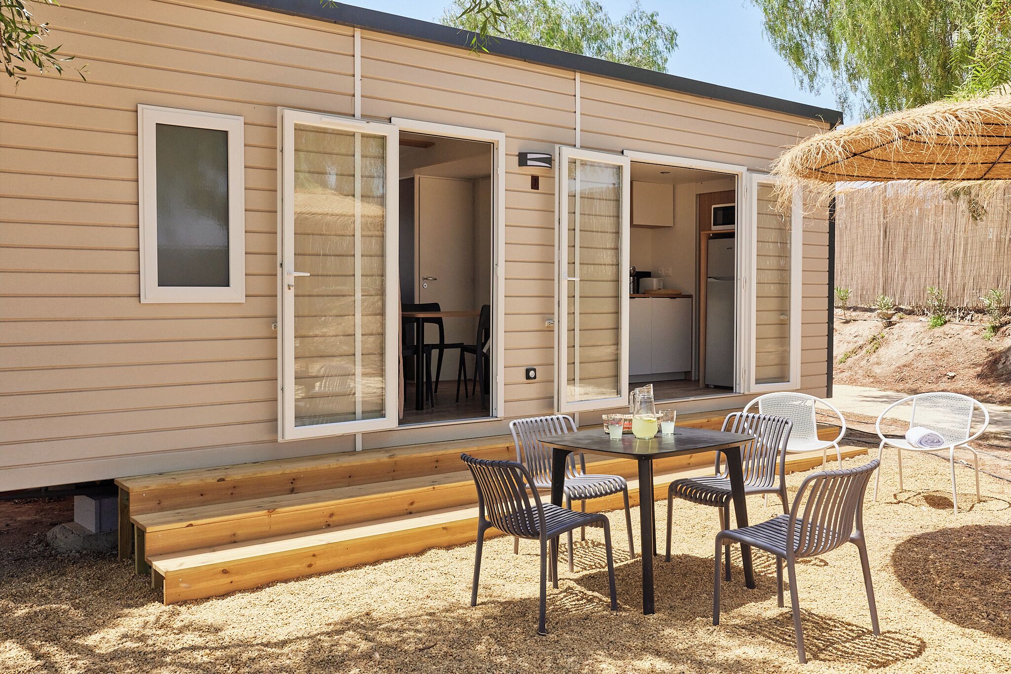 Een moderne stacaravan met een terras, compleet met een tafel, stoelen, een houten bank en een rieten parasol.