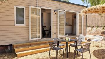 Een moderne stacaravan met een terras, compleet met een tafel, stoelen, een houten bank en een rieten parasol.