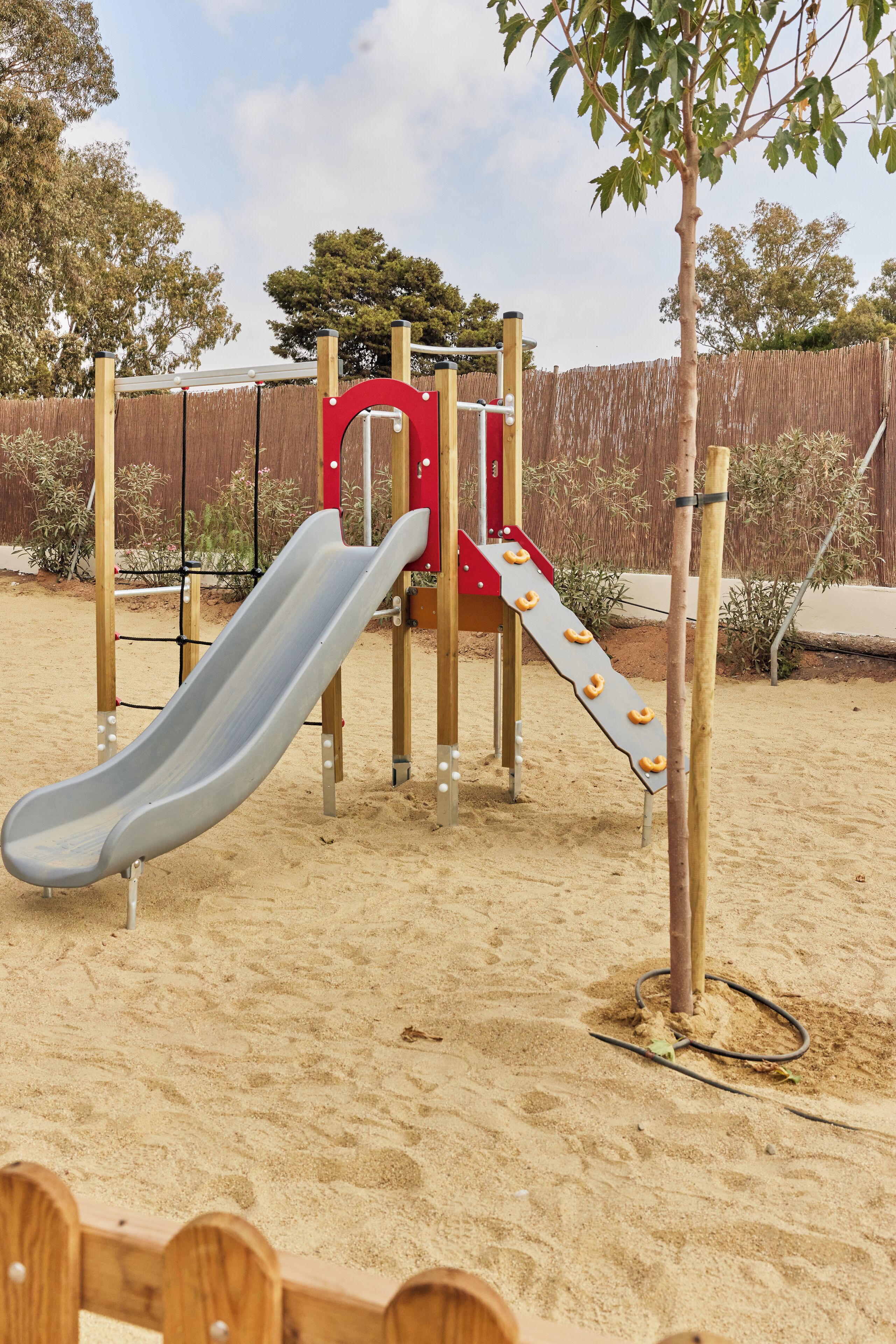 A playground with a slide and climbing structures, surrounded by sand and trees.