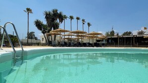 Piscina cristalina con sillas de descanso y sombrillas, rodeada de palmeras y un edificio con zona cubierta.