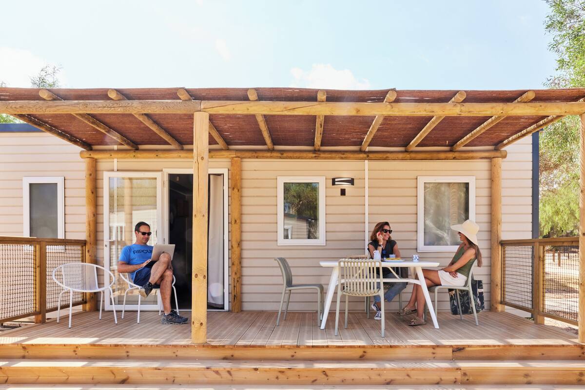 Eine Holzterrasse mit zwei Tischen und Stühlen, einem überdachten Bereich und einem Gebäude mit Fenstern und einer Tür.