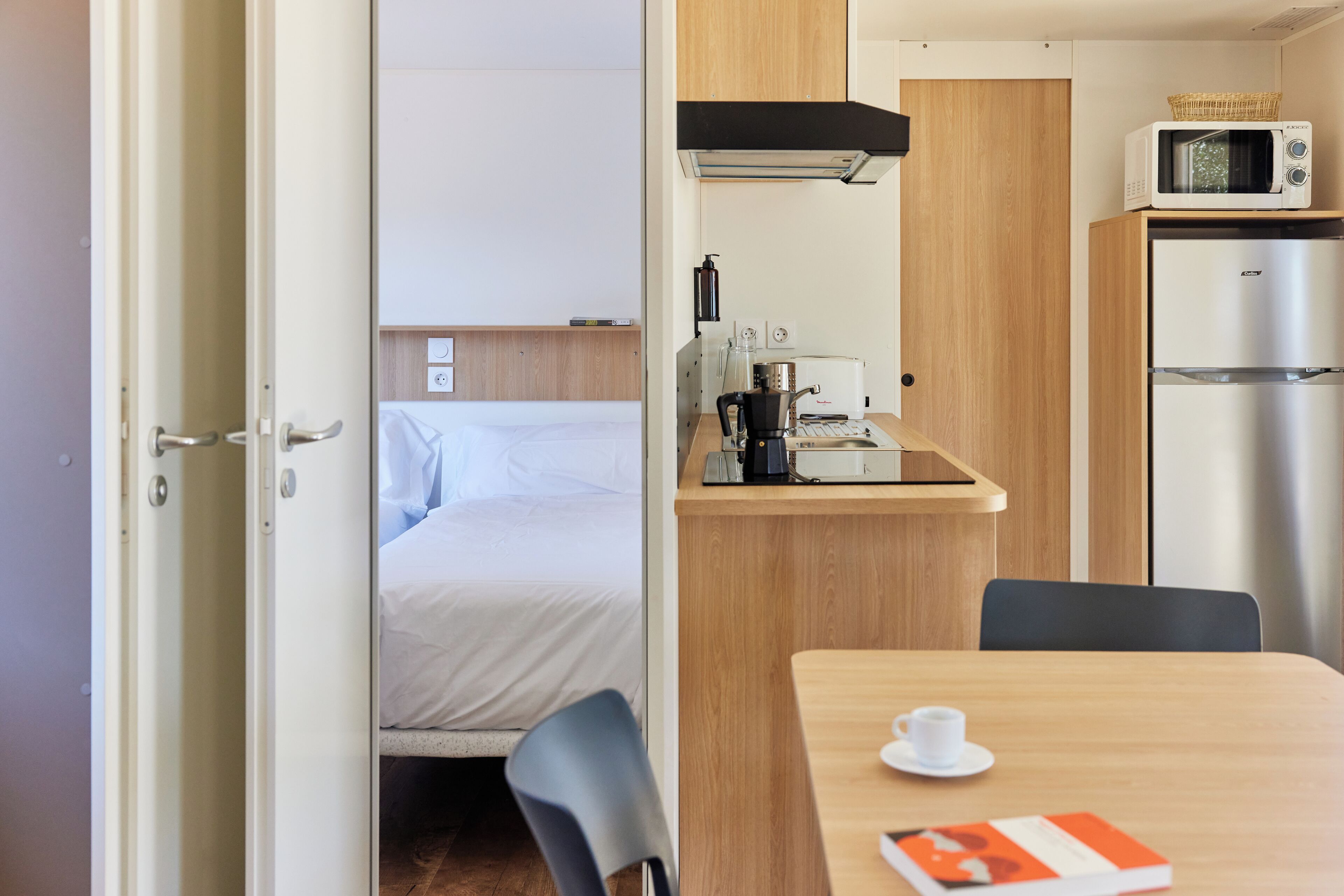 A compact room with a bed, a small kitchenette with a microwave and refrigerator, a table with a cup and a book, and a chair.