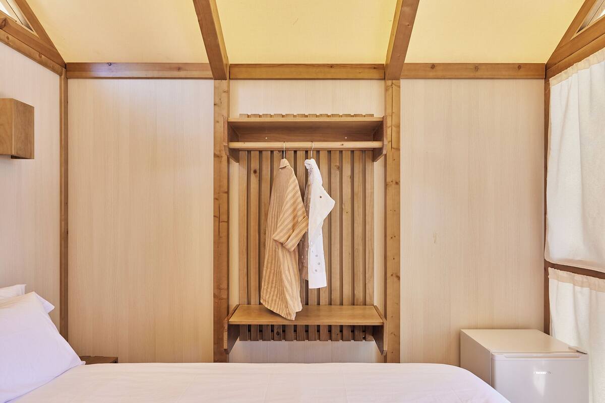 Ein Schlafzimmer mit einem Holzbett, einem Nachttisch, einem kleinen Regal und einem Kleiderhaken mit Kleidung.