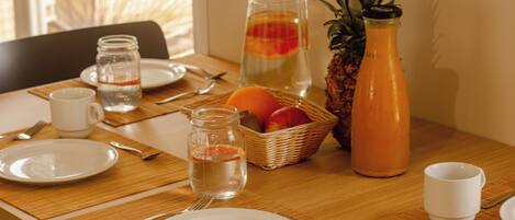 A breakfast table set with plates, cutlery, a pitcher of orange juice, a bottle of pineapple juice, a jar of jam, and a basket of fruit.
