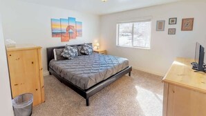 A bedroom with a bed, a desk, a window, and artwork on the walls. - Red Cliffs Condos - 6i (Moab)