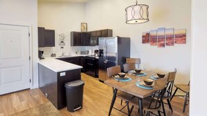 A modern kitchen with dark cabinets, a stainless steel refrigerator, and a dining area with a wooden table and chairs. - Red Cliffs Condos - 6i (Moab)