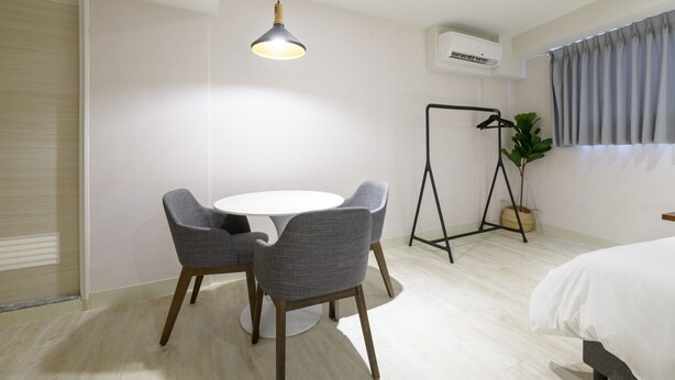 A modern hotel room with a round table, two grey chairs, a bed with white bedding, a hanging light fixture, an air conditioning unit, a window with grey curtains, and a potted plant. - Members Hotel at Taipei 101 (Taipei)