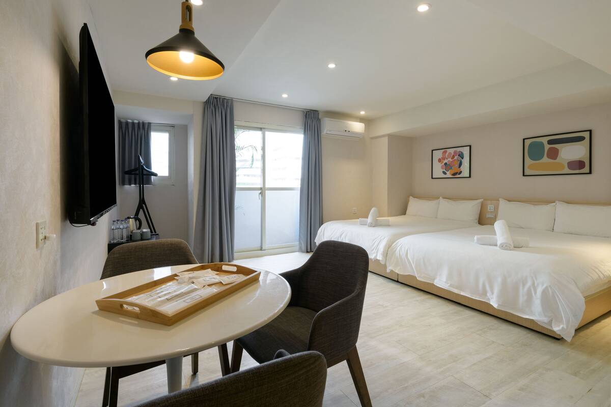 A hotel room with a bed, a desk with a tray, a chair, a TV, and a window with curtains.