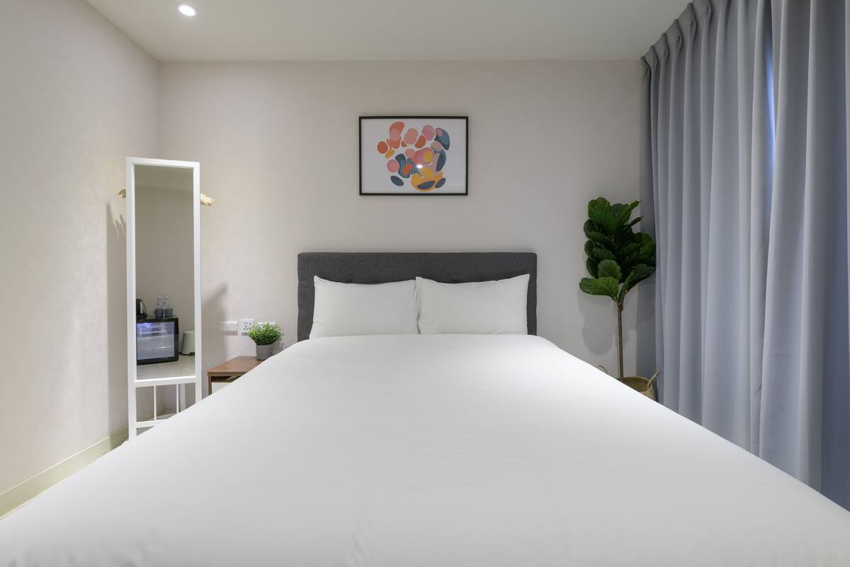 A hotel room with a large bed, a bedside table with a plant, a framed artwork on the wall, and a window with curtains.