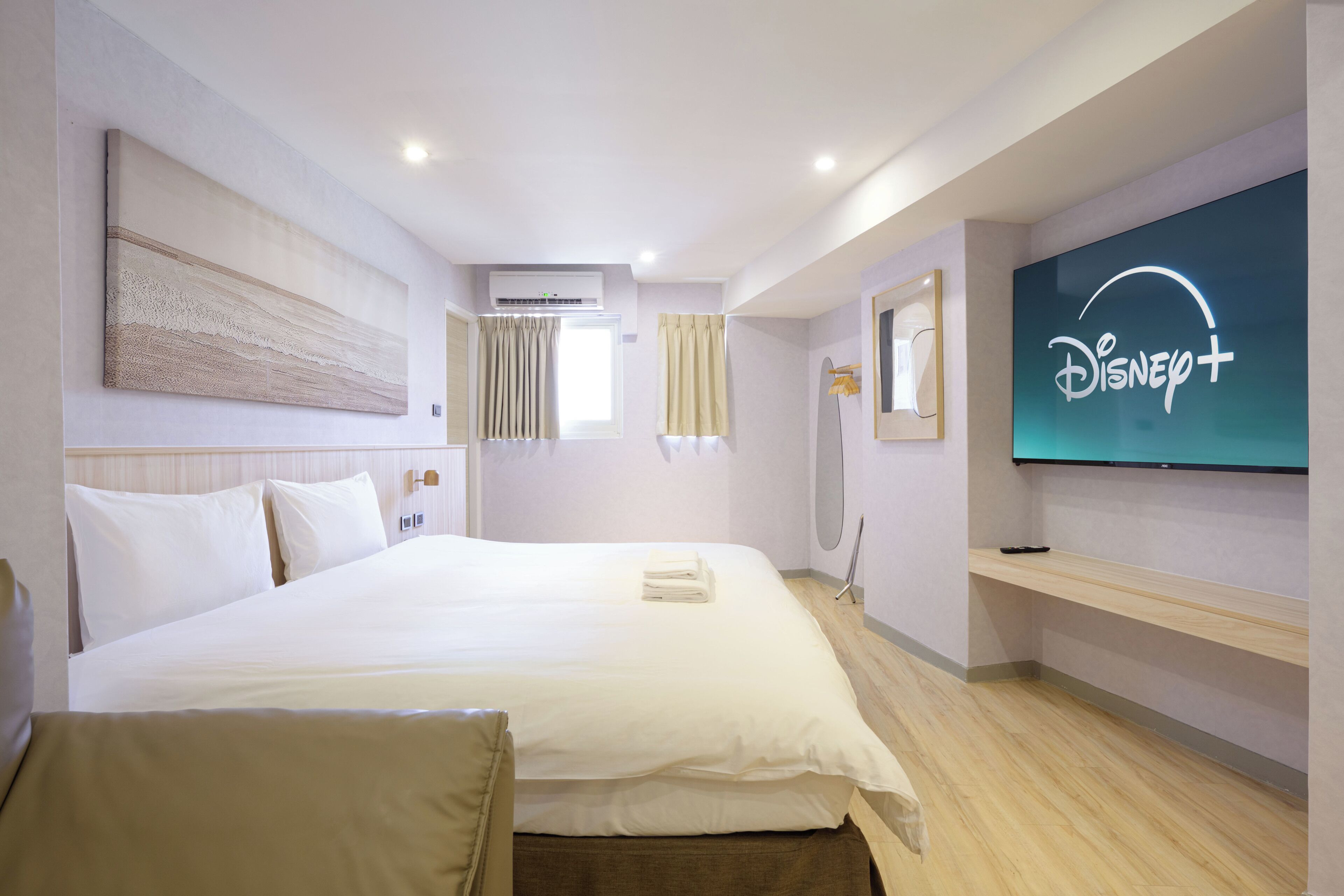 A hotel room with a large bed, a flat-screen TV mounted on the wall, a ceiling air conditioner, and a window with curtains.