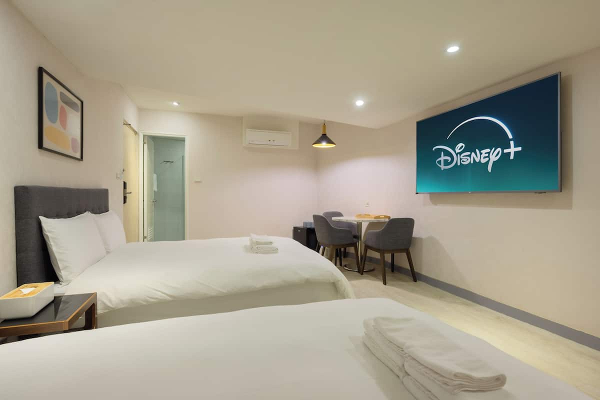A hotel room with two beds, a desk, and a chair. There is a Disney+ sign on the wall.