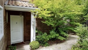 Una casa di legno con una porta bianca, un piccolo portico e un giardino con diverse piante e un vialetto sterrato.