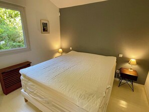 A bedroom with a large bed, two bedside lamps, a window with a view of greenery, and a small bedside table.