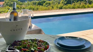 Mobiliario junto a la piscina para comer, con ensalada, queso, aceitunas y una botella de vino.