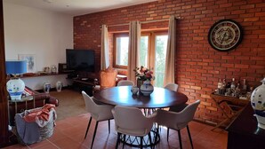 Un comedor con pared de ladrillo, una mesa redonda y sillas. Hay un televisor y un jarrón con flores sobre la mesa.