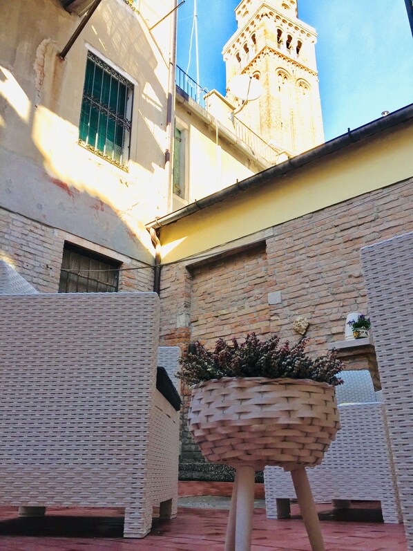 A courtyard with a brick wall, a hanging basket with plants, and a building with a tower in the background.
