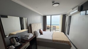 Une chambre d’hôtel avec un lit, un bureau, une bouilloire, des tasses et un miroir. La fenêtre offre une vue sur la plage.