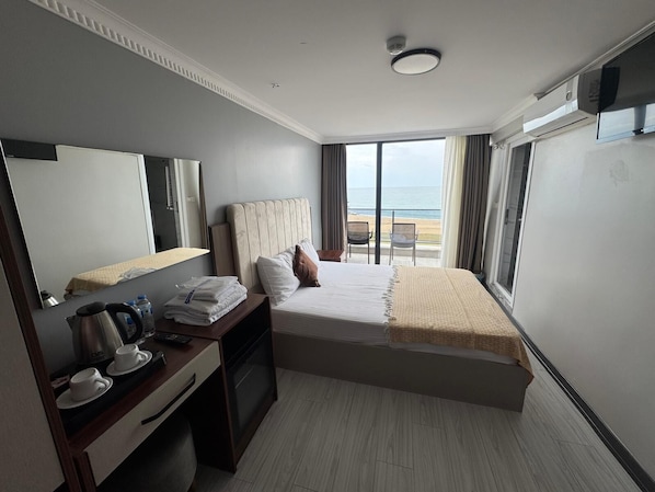 A hotel room with a bed, a desk with a kettle, cups, and a mirror. There is a view of the beach through the window. - Airport Kumsal Otel (Arnavutköy)