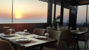 Un restaurant où les tables sont dressées pour le dîner, de grandes fenêtres offrant une vue sur le coucher de soleil et une mer calme en toile de fond.