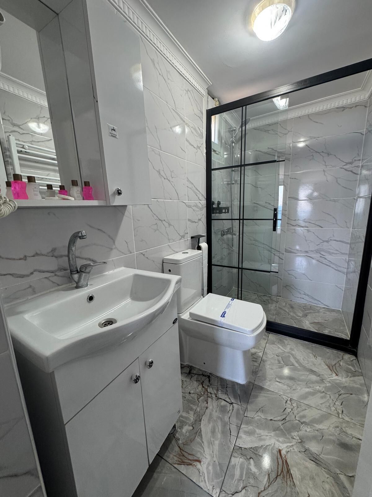 Un baño moderno con piso de mármol, un lavamanos blanco, inodoro y ducha con cerramiento de vidrio.