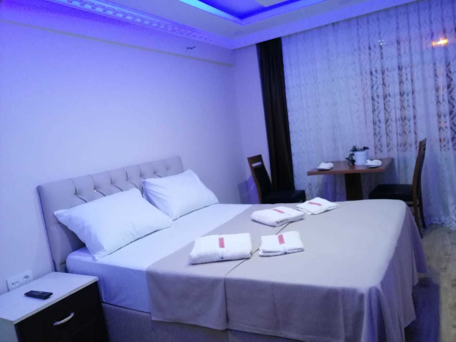 A neatly made bed with white linens and a grey headboard, a bedside table with a remote control, and a chair near a window with sheer curtains.