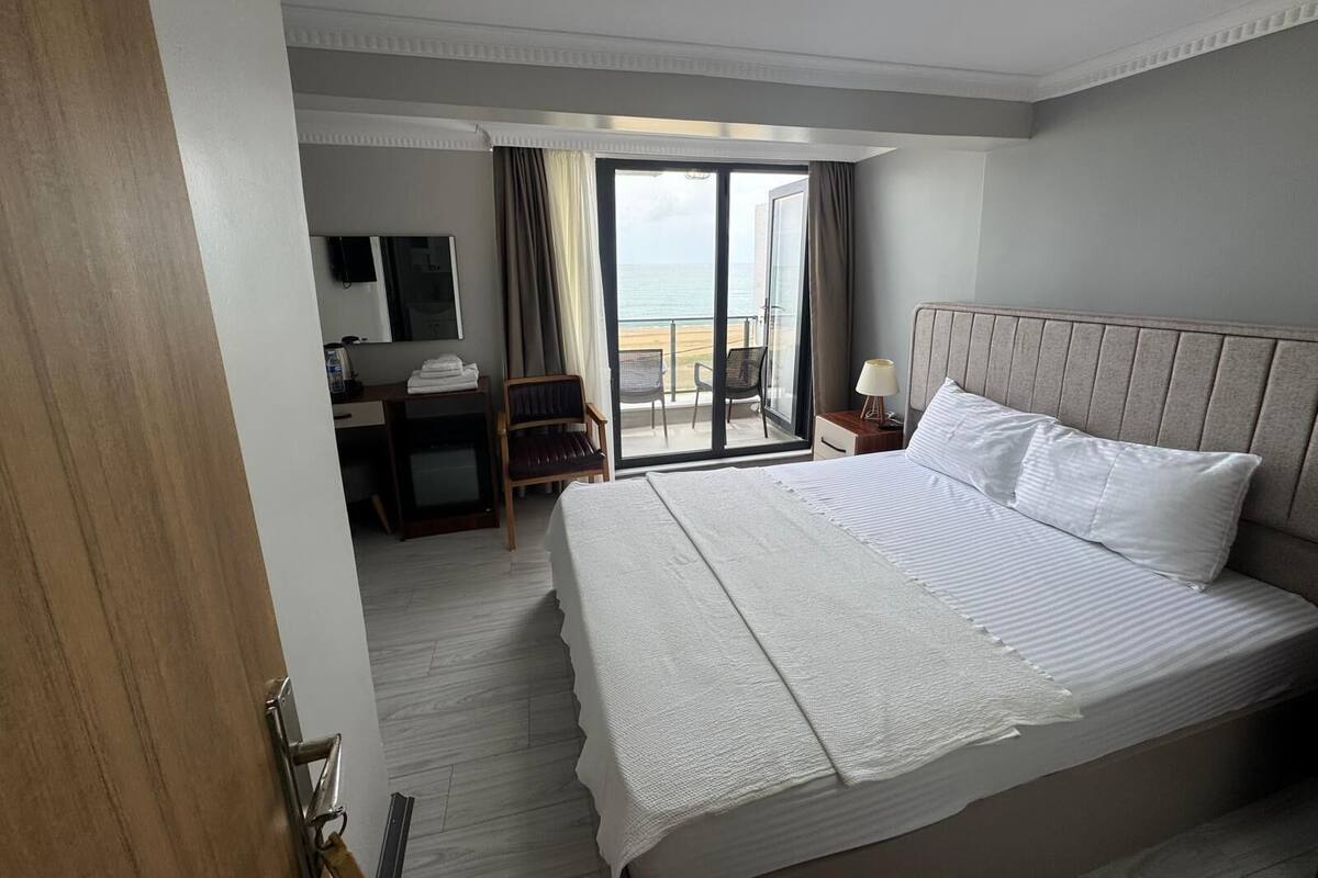 A hotel room with a large bed, a desk, a chair, and a view of the sea through a glass door.