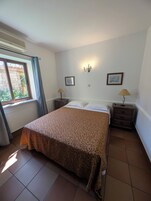 A bedroom with a bed, two bedside tables, a window with curtains, a wall-mounted light fixture, and a framed picture.