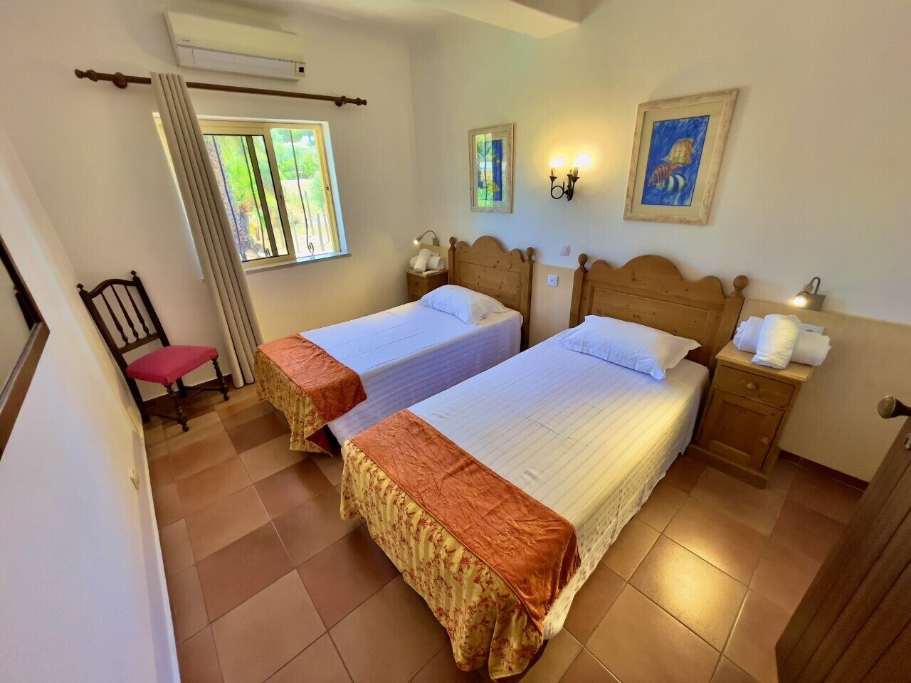 A bedroom with two beds, a window with curtains, a chair, a nightstand, and a wall-mounted lamp.