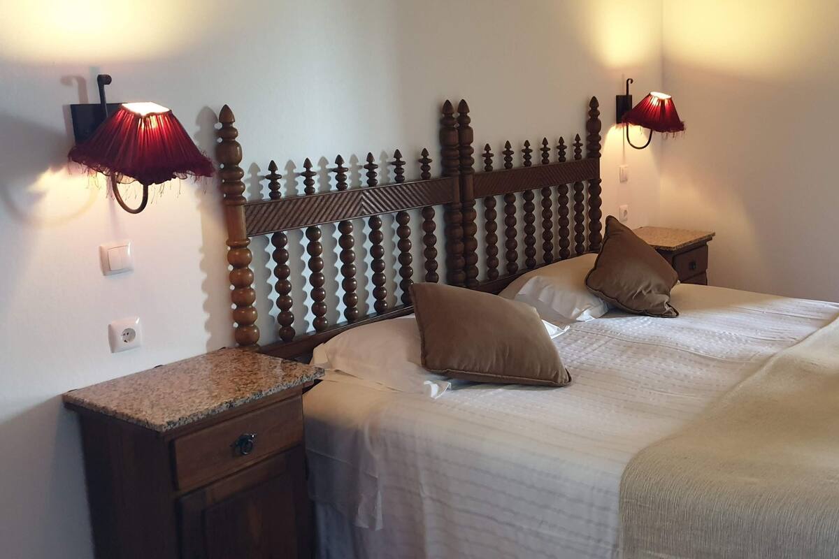 Un dormitorio con cabecera de madera, dos lámparas de noche, una cama con manta color beige y una mesita de noche con cajón.