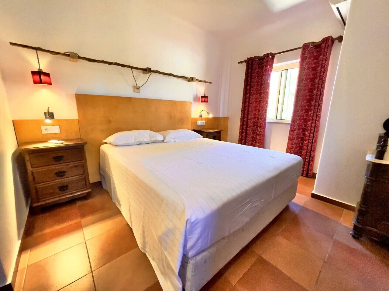Un dormitorio con una cama grande, un cabecero de madera, una mesita de noche con cajones, una lámpara y una ventana con cortinas rojas.