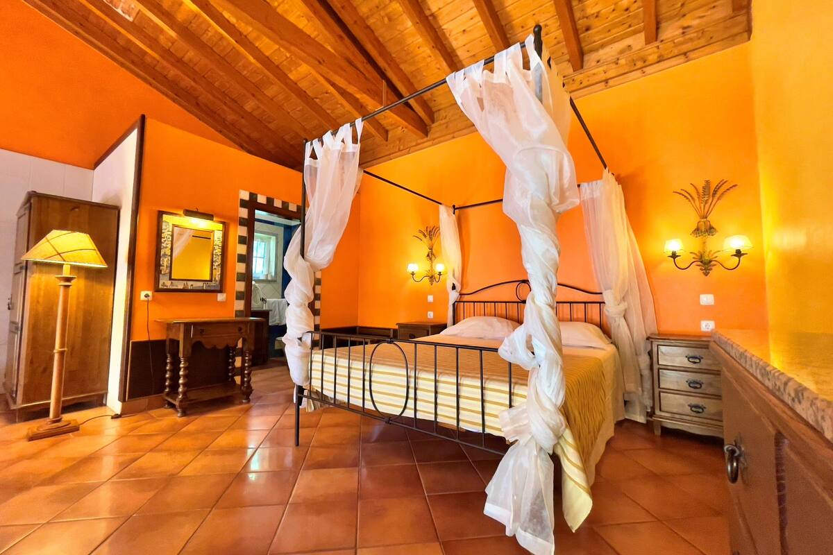 Una habitación con una cama con dosel, techo de madera, suelo de baldosas, una mesita de noche, una lámpara y un espejo.