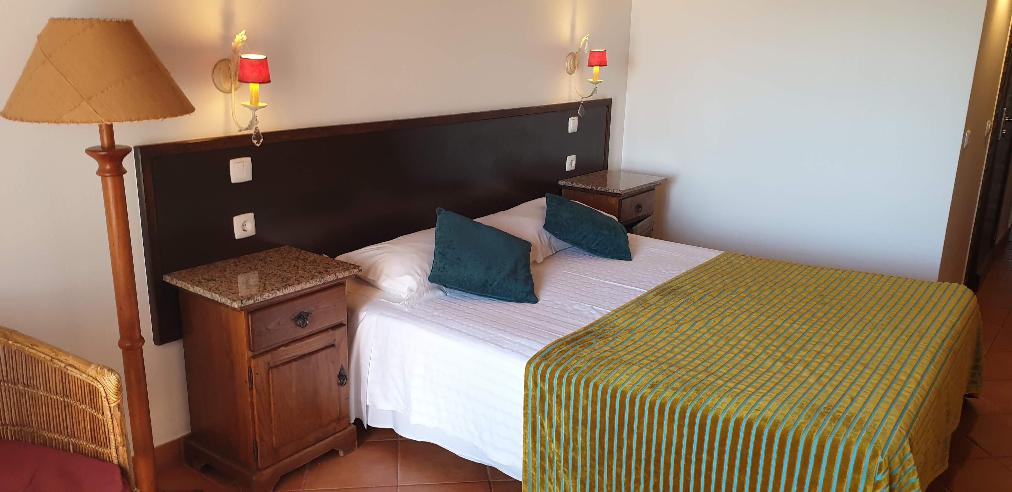 Una cama bien tendida con una colcha verde a rayas, dos almohadas, una mesita de noche de madera con una lámpara y un aplique de pared.