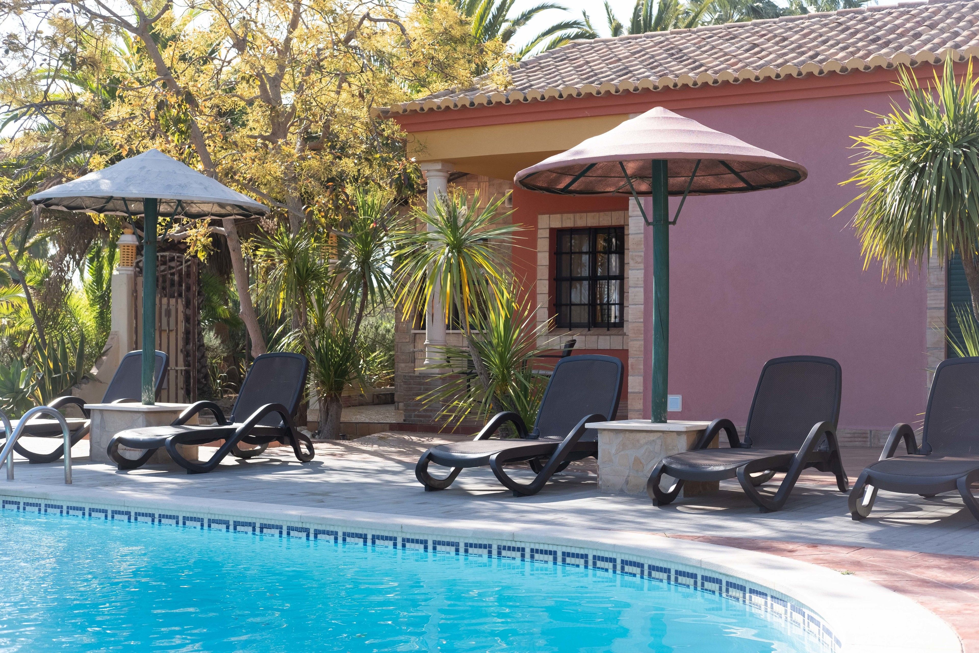 Un área junto a la piscina con sillones de descanso y sombrillas, un edificio con techo de tejas y vegetación tropical.