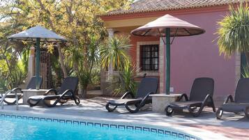 Un área junto a la piscina con sillones de descanso y sombrillas, un edificio con techo de tejas y vegetación tropical.