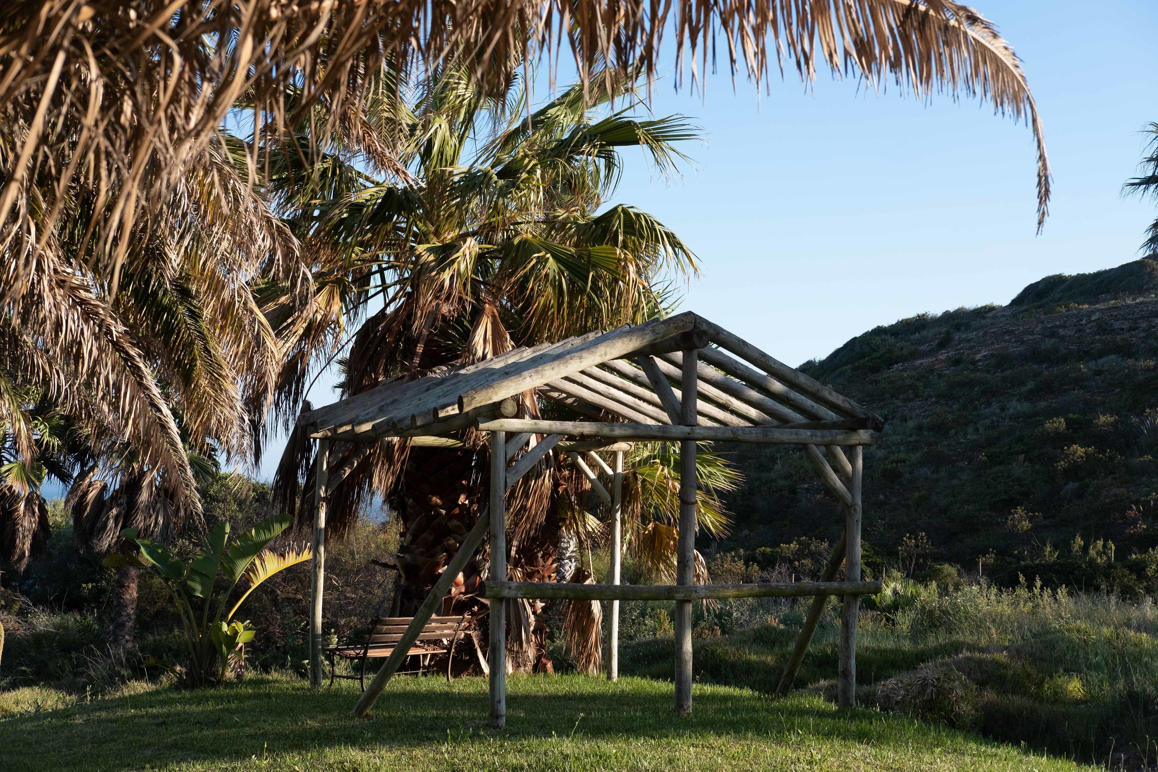 A wooden gazebo with a sloped roof, surrounded by palm trees and grass, set against a hilly backdrop.
