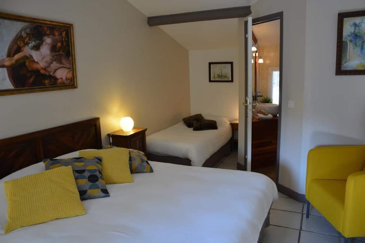 A hotel room with a large bed, two smaller beds, a yellow chair, and a painting on the wall.